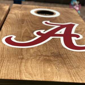 May include: Wooden cornhole board with a large crimson "A" logo outlined in white. The board has a natural wood grain finish and a circular hole for bean bag toss. A second board is partially visible.