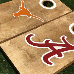 May include: Two wooden cornhole boards with team logos. One board features an orange longhorn logo with a white outline, and the other board has a crimson and white logo. Each board has a white-ringed hole.