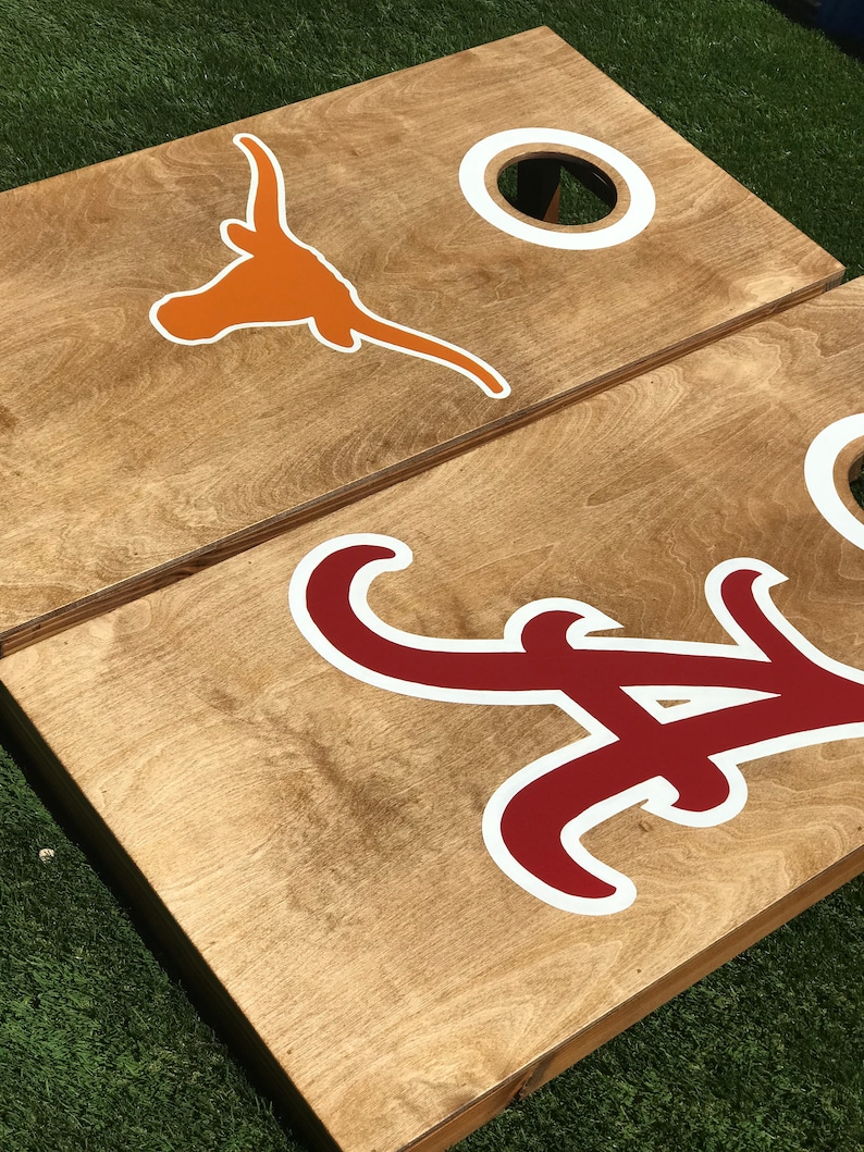 May include: Two wooden cornhole boards with team logos. One board features an orange longhorn logo with a white outline, and the other board has a crimson and white logo. Each board has a white-ringed hole.