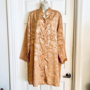 May include: A long, button-down robe in a golden-tan color, featuring a floral pattern. The robe has long sleeves and a mandarin collar. It is displayed on a white mannequin against a white door.