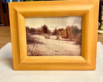 May include: A light-colored wooden picture frame with a photograph of a sandy beach scene. The photograph shows sand dunes, tall grasses, and a distant view of the ocean. The frame is rectangular with a simple, clean design.