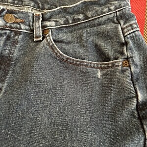 May include: Close-up of blue denim jeans. The jeans feature a button closure with the letters "R" and "CO" on the button. A small pocket is visible, edged with brown stitching.