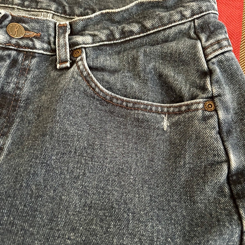 May include: Close-up of blue denim jeans. The jeans feature a button closure with the letters "R" and "CO" on the button. A small pocket is visible, edged with brown stitching.