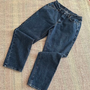 May include: Dark blue denim jeans. The jeans have a classic five-pocket design, a button and zip closure, and a straight leg cut. The jeans are laid flat on a textured surface.