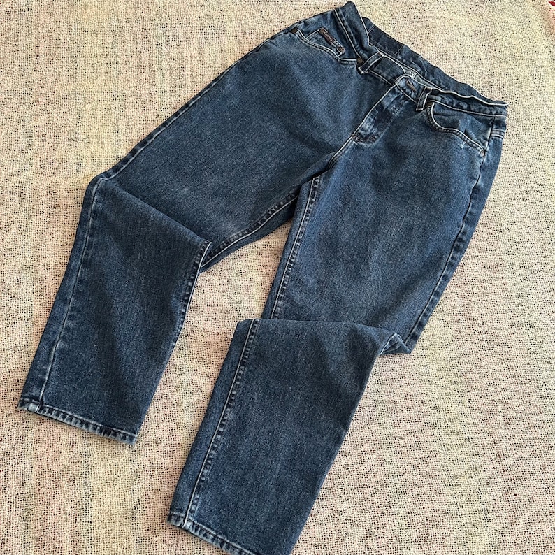 May include: Dark blue denim jeans. The jeans have a classic five-pocket design, a button and zip closure, and a straight leg cut. The jeans are laid flat on a textured surface.