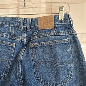 Puede incluir: Primer plano de unos vaqueros de mezclilla azules con un parche Lee de cuero marr&oacute;n en la parte trasera. Los vaqueros tienen dos bolsillos traseros, costuras amarillas y un dise&ntilde;o cl&aacute;sico. Los vaqueros est&aacute;n colgados de una percha de pl&aacute;stico transparente.