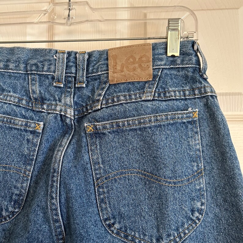 Puede incluir: Primer plano de unos vaqueros de mezclilla azules con un parche Lee de cuero marr&oacute;n en la parte trasera. Los vaqueros tienen dos bolsillos traseros, costuras amarillas y un dise&ntilde;o cl&aacute;sico. Los vaqueros est&aacute;n colgados de una percha de pl&aacute;stico transparente.