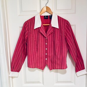 May include: A red and pink striped blouse with a white collar and cuffs. The long-sleeved shirt has a button-down front and is hanging on a wooden hanger. The blouse is made of a lightweight fabric.