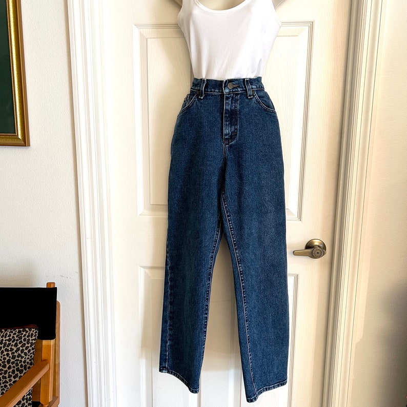May include: Dark wash blue jeans with a high waist and straight leg, combined with a white vest top. The jeans feature a classic five-pocket design and a button and zip fastening. The outfit is displayed on a white door.
