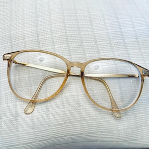 May include: A pair of eyeglasses with a translucent amber-colored frame and clear lenses. The temples are thin and metallic, featuring the word REGATTA printed on the inner side of the right temple. The frame has a rounded shape with metal hinges.