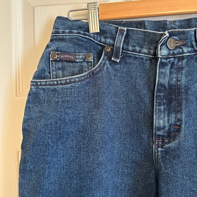 May include: Close-up of blue denim jeans with the brand name "RIDERS" on the front pocket. The jeans have a classic five-pocket design, a button and zip closure, and belt loops. The jeans are hanging on a wooden hanger.