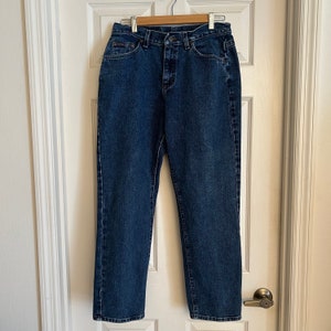 May include: A pair of blue denim jeans hanging on a wooden hanger. The jeans have a classic five-pocket design, a zip fly, and a button closure. The denim is a medium wash with subtle fading.