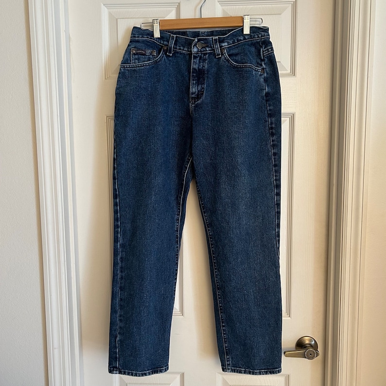 May include: A pair of blue denim jeans hanging on a wooden hanger. The jeans have a classic five-pocket design, a zip fly, and a button closure. The denim is a medium wash with subtle fading.