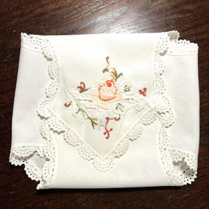 May include: White handkerchief with a floral embroidery design. The embroidery features an orange rose, green leaves, and small blue and red accents. The edges are trimmed with a delicate white lace.