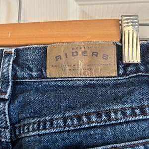 May include: Close-up of blue denim jeans with a brown leather patch. The patch reads "DENIM RIDERS" with additional text: "BASIC COMFORTABLE SATISFACTION AUTHENTIC SUPERIOR FIT GUARANTEED". The jeans are hanging on a wooden hanger.