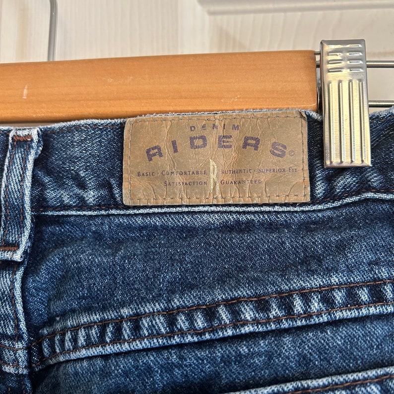 May include: Close-up of blue denim jeans with a brown leather patch. The patch reads "DENIM RIDERS" with additional text: "BASIC COMFORTABLE SATISFACTION AUTHENTIC SUPERIOR FIT GUARANTEED". The jeans are hanging on a wooden hanger.