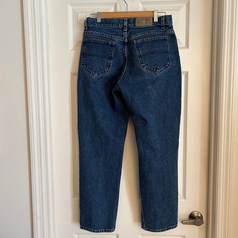 May include: Dark wash denim jeans hanging on a wooden hanger. The jeans have a classic five-pocket design with a straight leg cut. The back pockets have a curved stitch detail. A leather patch is visible at the waistband.