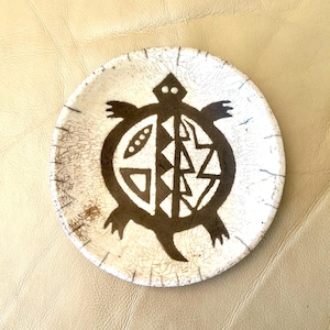 May include: A small, round, ceramic plate with a brown turtle design. The turtle is stylized with geometric shapes and is centered on a white background with a crackle finish. The plate has a slightly raised edge.