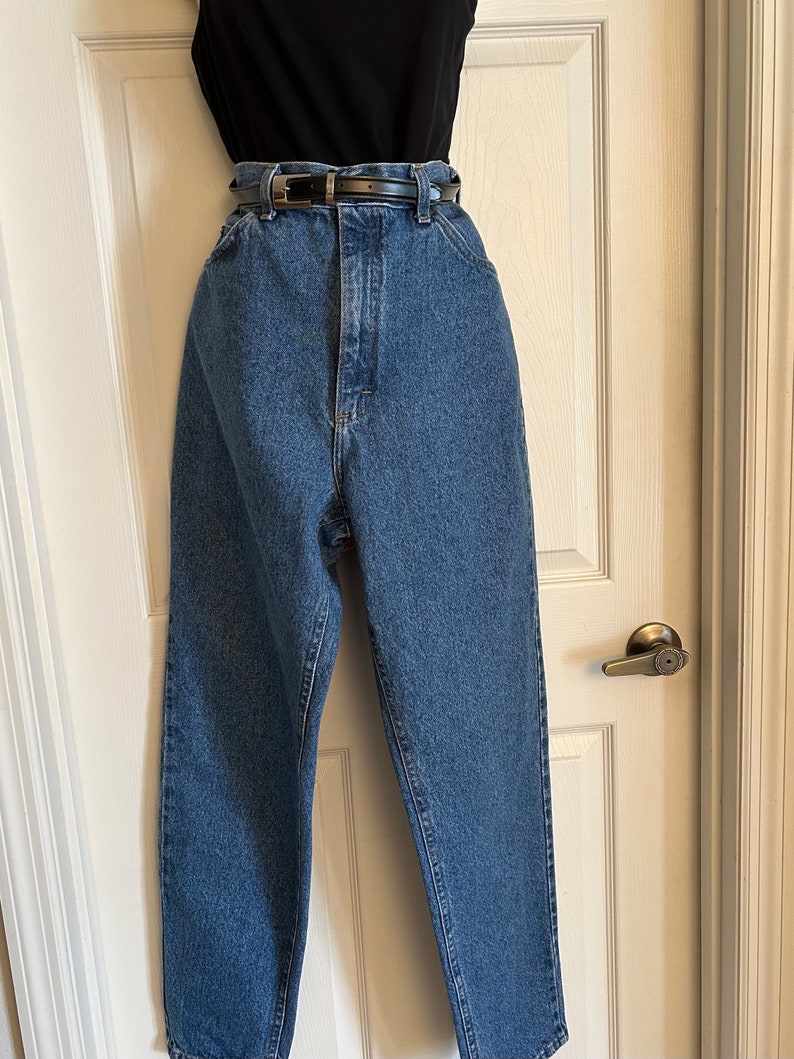 Puede incluir: Vaqueros de mezclilla azul de lavado medio, de cintura alta, con cintur&oacute;n de cuero negro. Los vaqueros tienen un dise&ntilde;o cl&aacute;sico de cinco bolsillos y una pernera recta. Una camiseta negra es visible encima de los vaqueros. Los vaqueros se muestran en una puerta blanca.