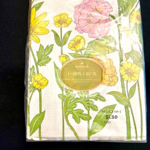 May include: A Hallmark table cover featuring a floral design with yellow and pink flowers, green leaves, and stems on a white background. The package reads "60 in. x 102 in." and "Table Cover."