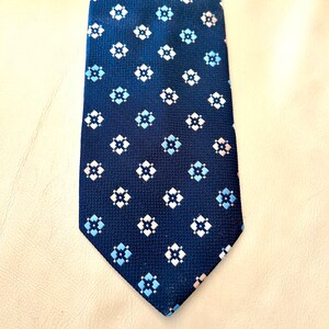 May include: Navy blue tie with a repeating pattern of small, four-petal floral designs in white, light blue, and pale pink. The tie has a textured surface and a classic pointed end.