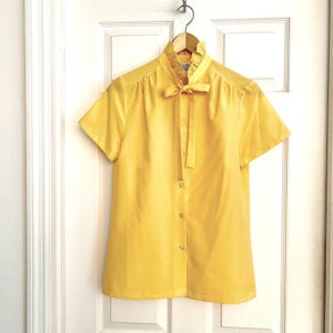May include: A short-sleeved, bright yellow blouse with a high, ruffled collar and a bow tie. The blouse has a button-down front and is hanging on a wooden hanger against a white door.