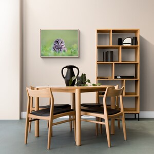 May include: A round wooden dining table with four chairs. The table is set with a black vase and a plant. A framed print of an owl is hanging on the wall behind the table. A wooden bookshelf with black accents is in the background.