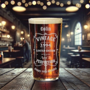 May include: A clear glass pint glass with a brown liquid inside. The glass has an etched design that says "Colin Vintage 1994 Limited Edition Aged to Perfection Happy 30th Birthday".