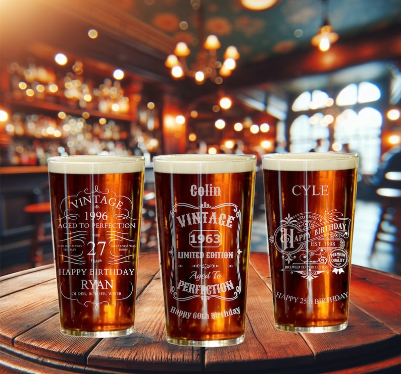 May include: Three personalized pint glasses with etched designs celebrating birthdays. The glasses are filled with a dark amber liquid and are sitting on a wooden table. The text on the glasses reads "Vintage 1996 Aged to Perfection Happy 27th Birthday Ryan", "Colin Vintage 1963 Limited Edition Aged to Perfection Happy 60th Birthday", and "Cyle Happy 25th Birthday".