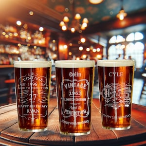 May include: Three personalised pint glasses with etched designs celebrating birthdays. The glasses are filled with a dark amber liquid and are sitting on a wooden table. The text on the glasses reads "Vintage 1996 Aged to Perfection Happy 27th Birthday Ryan", "Colin Vintage 1963 Limited Edition Aged to Perfection Happy 60th Birthday", and "Cyle Happy 25th Birthday".