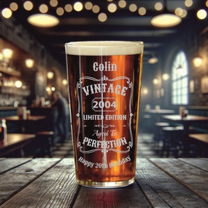 May include: A clear glass pint glass with a brown liquid inside. The glass has a white design that says "Colin Vintage 2004 Limited Edition Aged to Perfection Happy 20th Birthday".