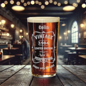 May include: A clear glass pint glass with a brown liquid inside. The glass has an etched design that says "Colin Vintage 1984 Limited Edition Aged to Perfection Happy 40th Birthday".