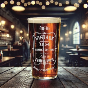 May include: A clear glass pint glass with a brown liquid inside. The glass has an etched design that says "Colin Vintage 1954 Limited Edition Aged to Perfection Happy 70th Birthday".