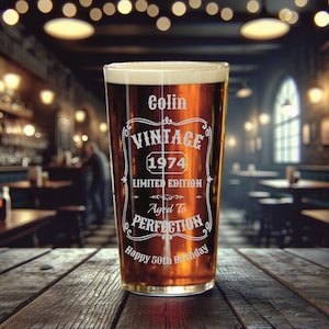 May include: A clear glass pint glass with a brown liquid inside. The glass has an etched design that says "Colin Vintage 1974 Limited Edition Aged To Perfection Happy 50th Birthday".