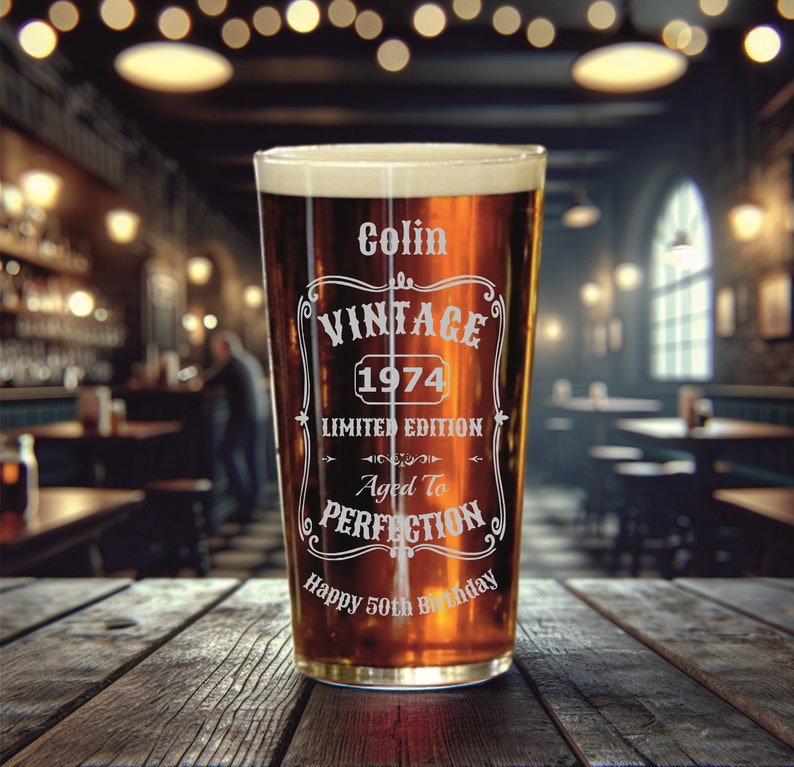 May include: A clear glass pint glass with a brown liquid inside. The glass has an etched design that says "Colin Vintage 1974 Limited Edition Aged To Perfection Happy 50th Birthday".