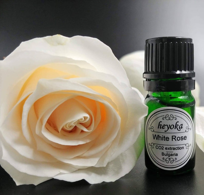 May include: A small green glass bottle with a black lid containing white rose essential oil. The label reads "heyoka White Rose CO2 extraction Bulgaria". A white rose is in the background.