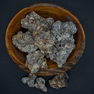 May include: A wooden bowl filled with several pieces of dark, textured resin. The resin pieces have a mix of brown, gray, and white hues. The bowl is made of light brown wood and sits on a dark surface.