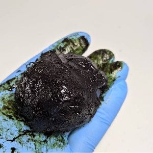 May include: A gloved hand holds a dark brown, almost black, sticky substance. The substance is textured and appears to be a thick liquid or paste.