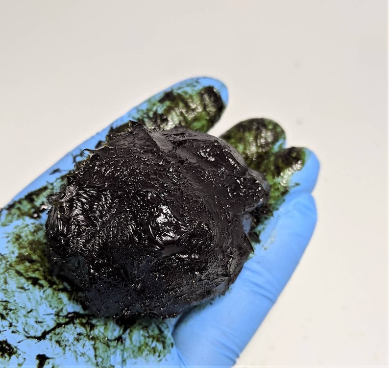 May include: A gloved hand holds a dark brown, almost black, sticky substance. The substance is textured and appears to be a thick liquid or paste.
