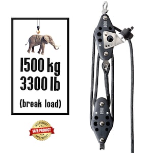 May include: A black pulley system with two black pulleys and a silver hook. The pulley system is attached to a black rope. The rope is attached to a white sign with black text that reads "1500 kg 3300 lb (break load)". A red and gold sticker with the text "SAFE PRODUCT" is at the bottom of the sign.