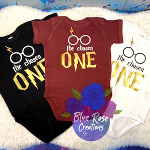 May include: Three baby onesies in black, burgundy, and white, each with a gold graphic of glasses, a lightning bolt, and the words "the chosen ONE." The onesies are decorated with a floral design and the text "Blue Rose Creations."