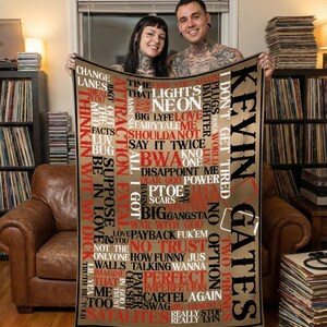 May include: A tan blanket featuring the name "Kevin Gates" in large black letters. The blanket is covered in various words and phrases in red and white, with a brown border. The blanket is being held by two people.