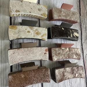 Puede incluir: Una colección de ocho pinzas para el pelo rectangulares. Cada pinza presenta un patrón diferente de piel de vaca en tonos marrón, beige y crema. Las pinzas tienen cierres de varios colores, incluyendo crema, marrón y malva.