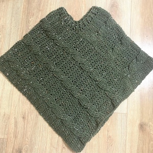 May include: A dark green, hand-knitted poncho with a cable knit design. The poncho is square-shaped with a v-neck opening. The yarn has flecks of white and other colors throughout. The texture of the knit is visible.