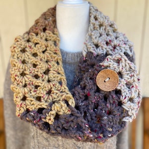 May include: A crocheted infinity scarf in brown, beige, and cream with a wooden button. The scarf is made with a textured stitch and has a speckled yarn.