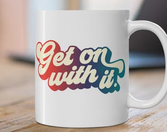 May include: White ceramic mug with the phrase "Get on with it" in a retro, colorful design. The text is in red, orange, blue, and purple. The mug has a curved handle and is on a wooden surface.