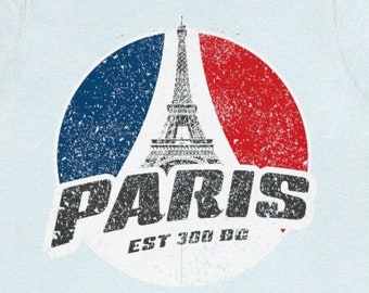 May include: A light blue t-shirt with a distressed graphic of the Eiffel Tower and the text "PARIS EST 300 BC" in a circle with red, white, and blue accents.