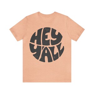 Pu&ograve; includere: Una t-shirt color pesca con un grafico circolare nero che dice "Hey Y'all" in un font retr&ograve;.