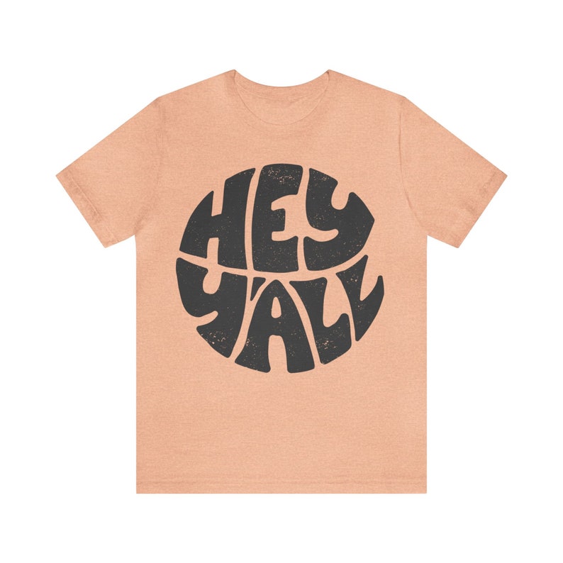 Pu&ograve; includere: Una t-shirt color pesca con un grafico circolare nero che dice "Hey Y'all" in un font retr&ograve;.