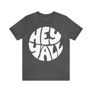 Pu&ograve; includere: Una maglietta grigio scuro con una grafica circolare bianca che dice "Hey Y'all" in un font retr&ograve;.
